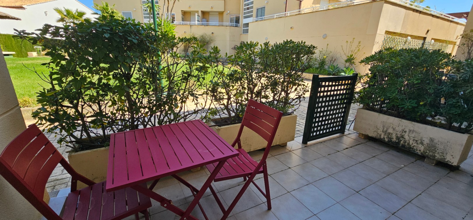 Terraza privada con suelo de baldosas, mobiliario de madera en color granate y jardineras con setos que dan acceso directo a las zonas comunes de la urbanización.