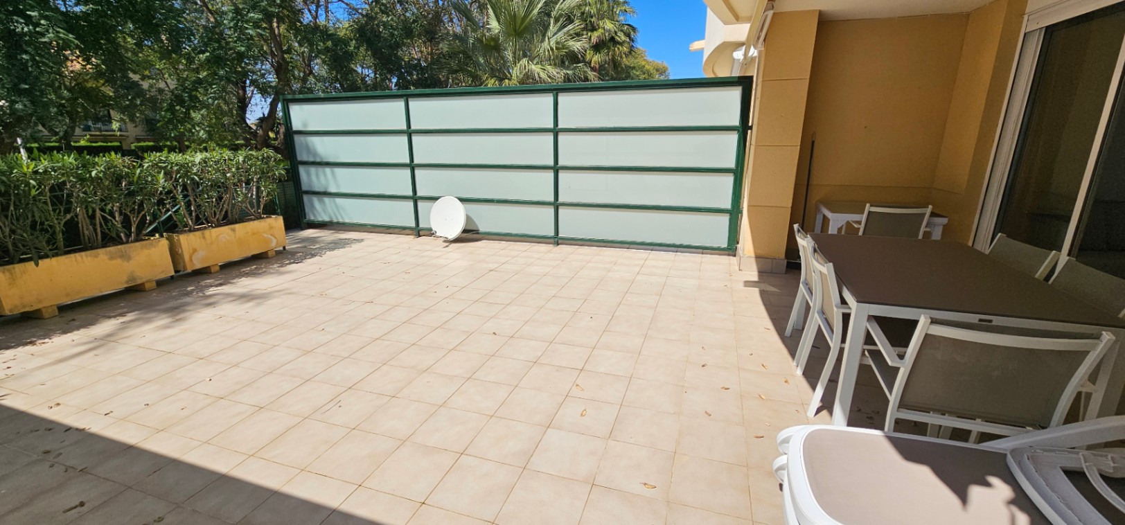 Terraza privada de orientación sur con suelo cerámico, zona de comedor exterior y paneles de privacidad, rodeada de vegetación y árboles maduros.