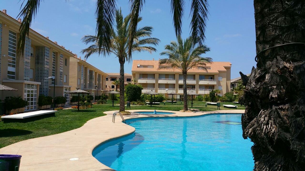 Zona comunitaria con piscina de diseño curvo rodeada de césped, palmeras y sombrillas, situada frente a los bloques de apartamentos de la urbanización.