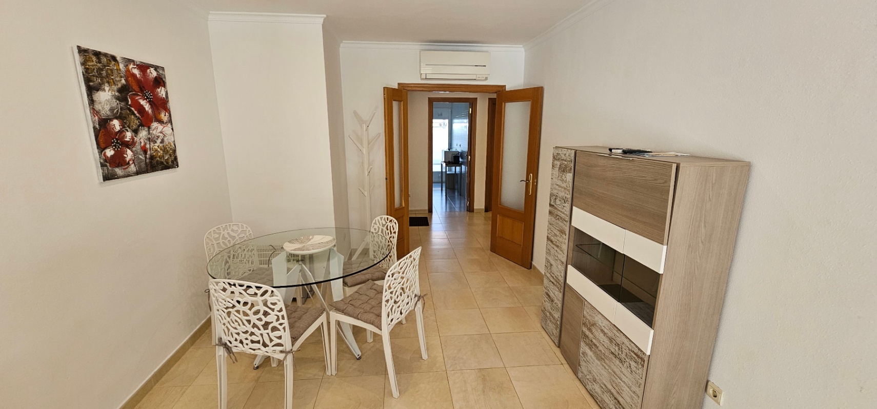Zona de comedor con mesa circular de cristal, sillas de diseño blanco y aparador moderno, conectada mediante puertas dobles de madera al resto de la vivienda.
