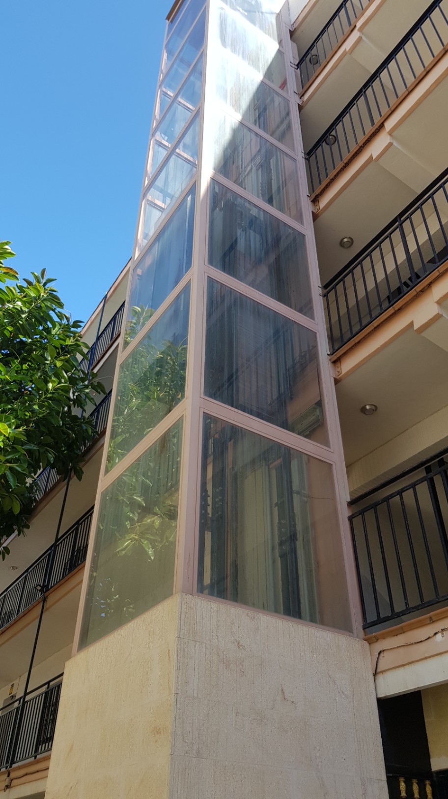 Ascensor panorámico exterior de cristal con base de piedra, integrado en la fachada del edificio residencial junto a los balcones con barandillas negras.