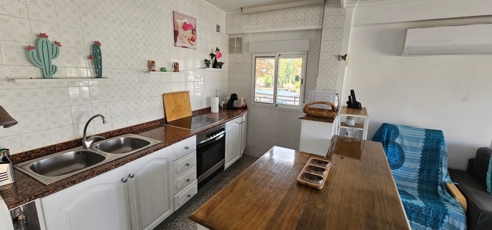 Cocina de concepto abierto con encimera de granito, fregadero doble de acero inoxidable y mesa de madera, integrada en un espacio luminoso con aire acondicionado.