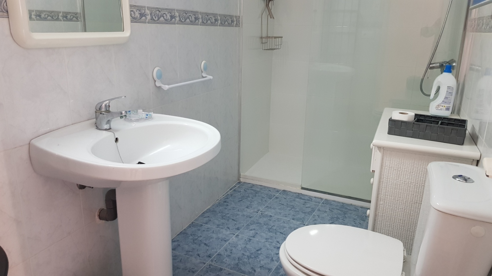 Cuarto de baño con suelo de gres azul, lavabo de pedestal y ducha con mampara de cristal. Incluye azulejos blancos con cenefa decorativa y mobiliario auxiliar.
