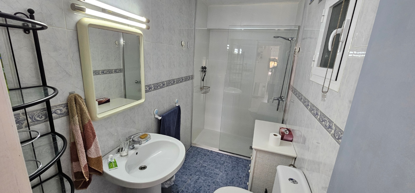 Cuarto de baño funcional con ducha de obra, mampara de cristal, lavabo de pedestal y suelos cerámicos azules que contrastan con el alicatado clásico.