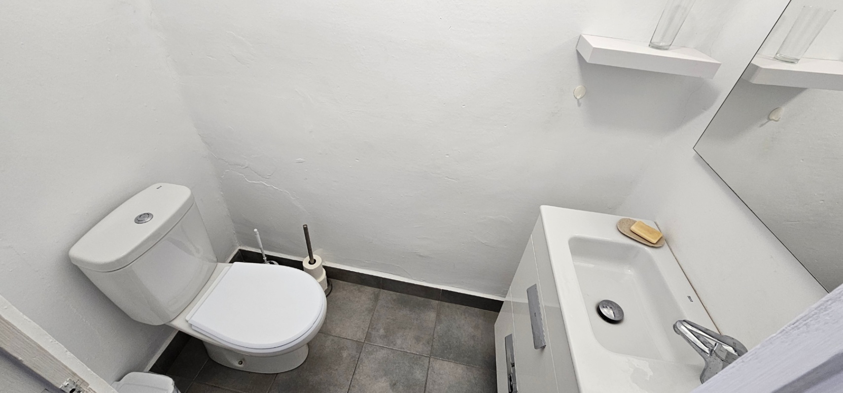 Cuarto de baño funcional con inodoro blanco, lavabo integrado en mueble de almacenaje y suelo de baldosas grises. Incluye un espejo y estante auxiliar sobre paredes blancas.