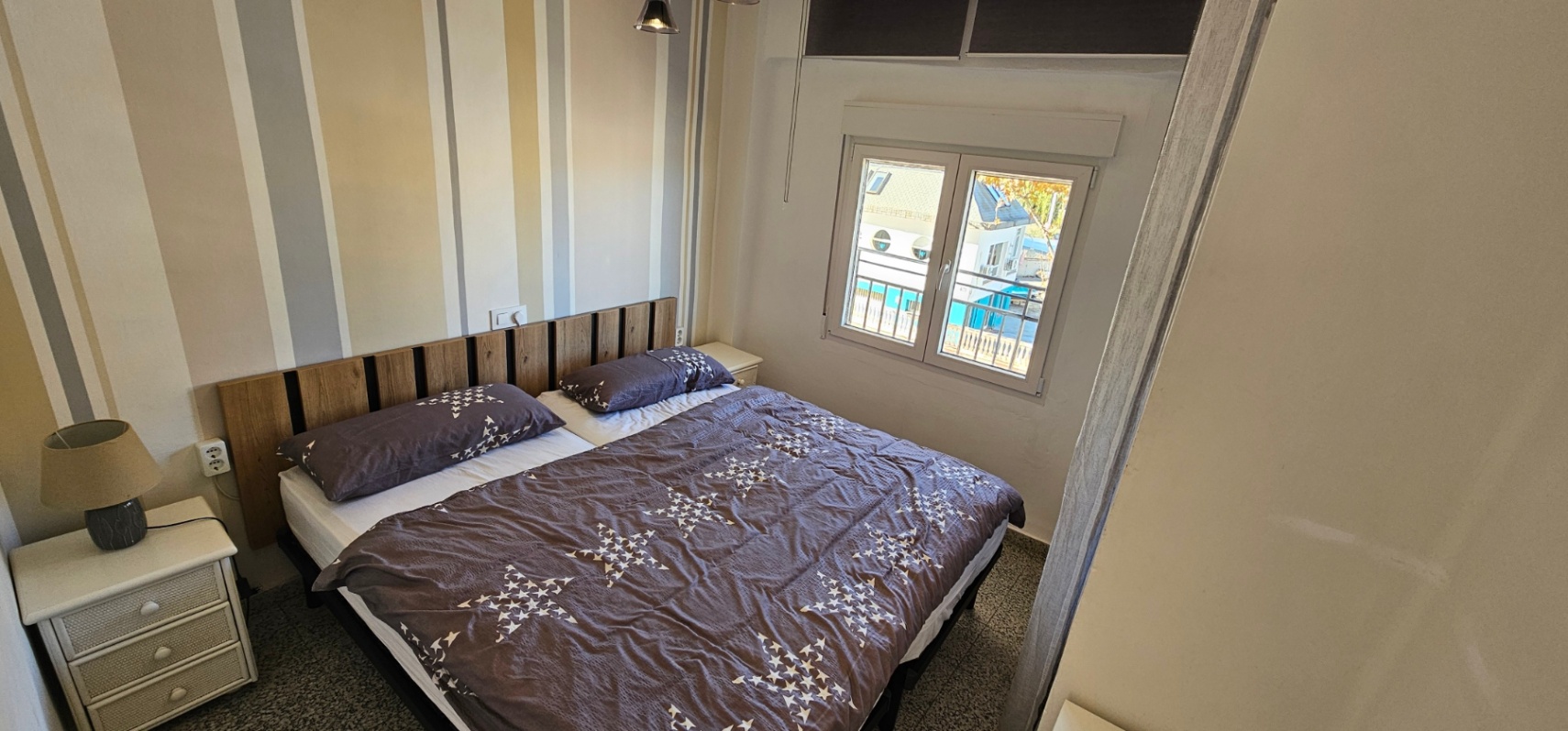Dormitorio con cama doble, cabecero de listones de madera y papel pintado a rayas. La ventana exterior ofrece luz natural y vistas despejadas hacia la zona de la marina.