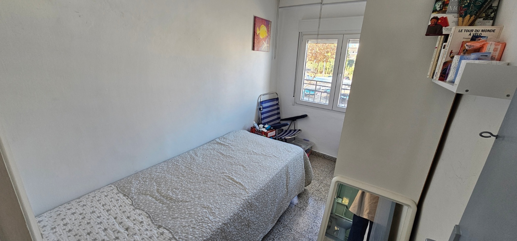 Dormitorio individual con cama de 90 cm, suelos de terrazo original y ventana exterior que aporta luz natural. Incluye estantería integrada y paredes en tono blanco.