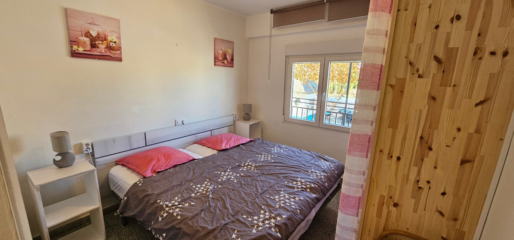 Dormitorio luminoso con cama doble, cabecero de madera clara y mesitas auxiliares blancas. Incluye ventana exterior con persiana y vistas despejadas al entorno.