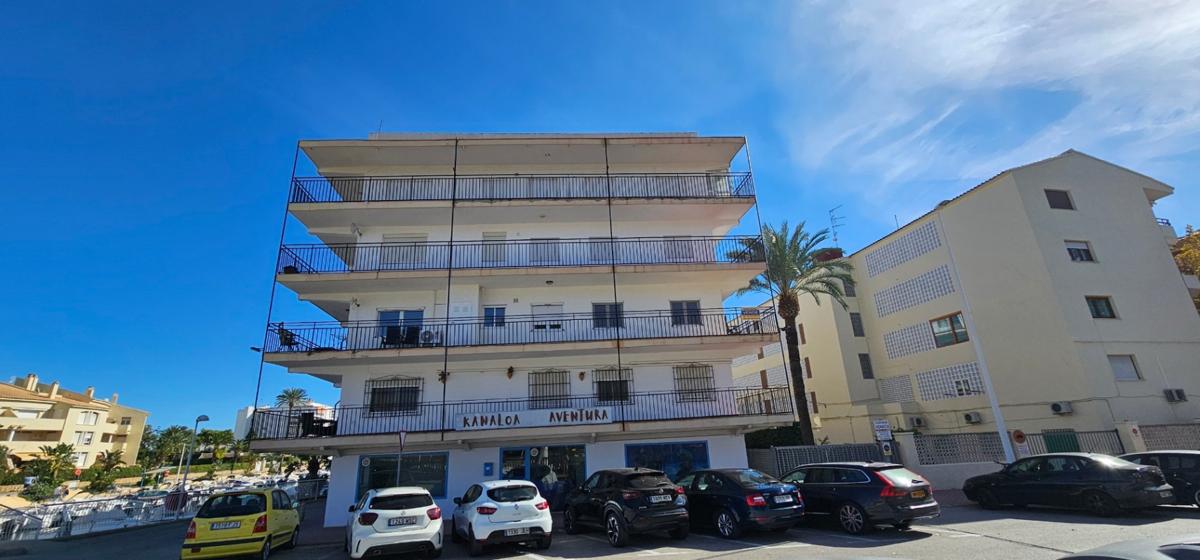 Fachada exterior del edificio con amplias terrazas perimetrales y barandillas metálicas, situado junto a la zona de aparcamiento y a pocos pasos de la marina.