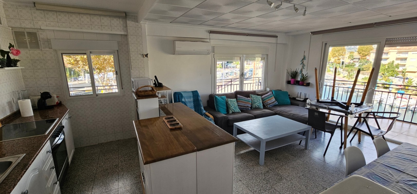 Salón-comedor de planta abierta con cocina integrada, barra de madera y acceso directo a una terraza con vistas al canal y los barcos de Jávea.