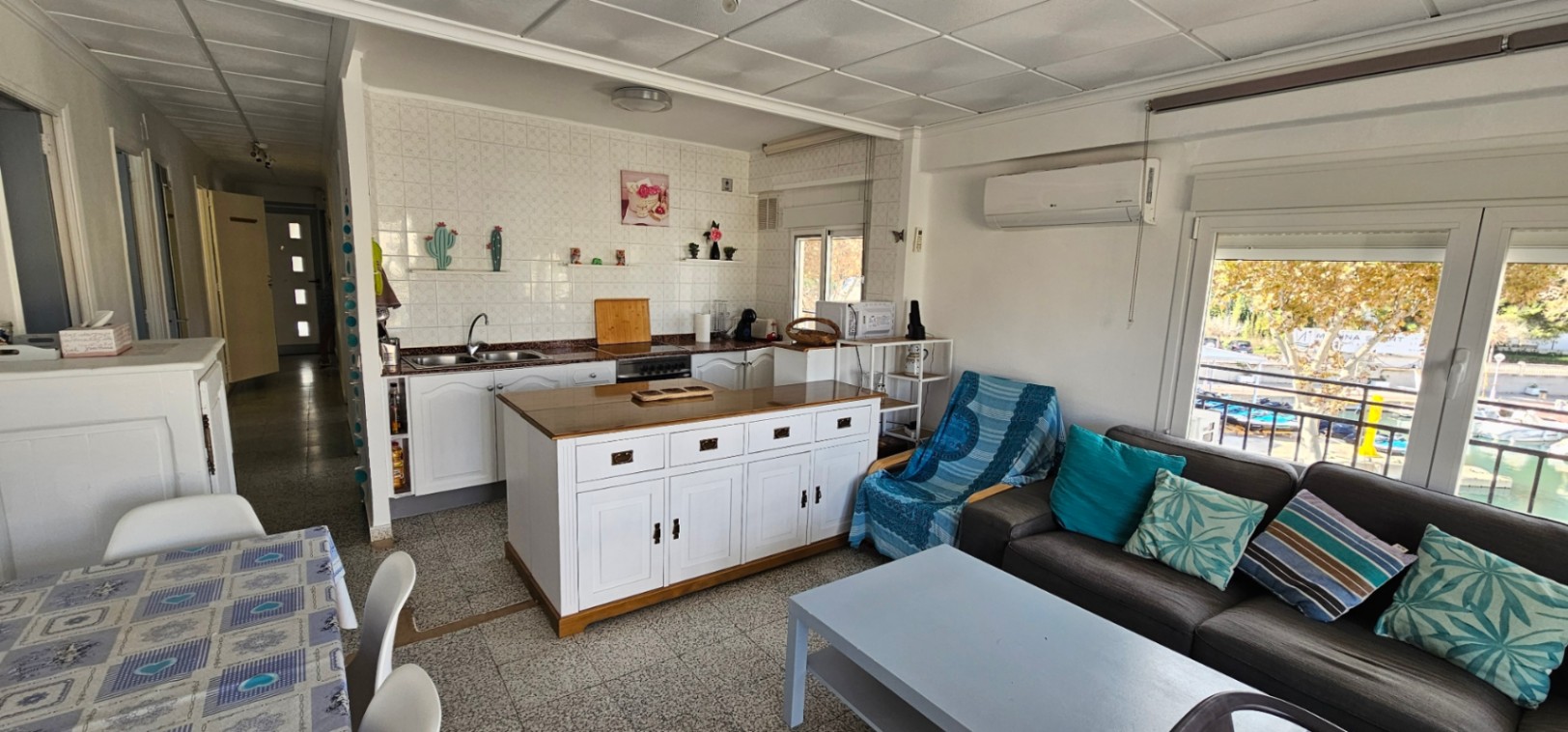 Salón-comedor de planta abierta con cocina integrada, isla central blanca y acceso directo a la terraza con vistas al canal y las embarcaciones.