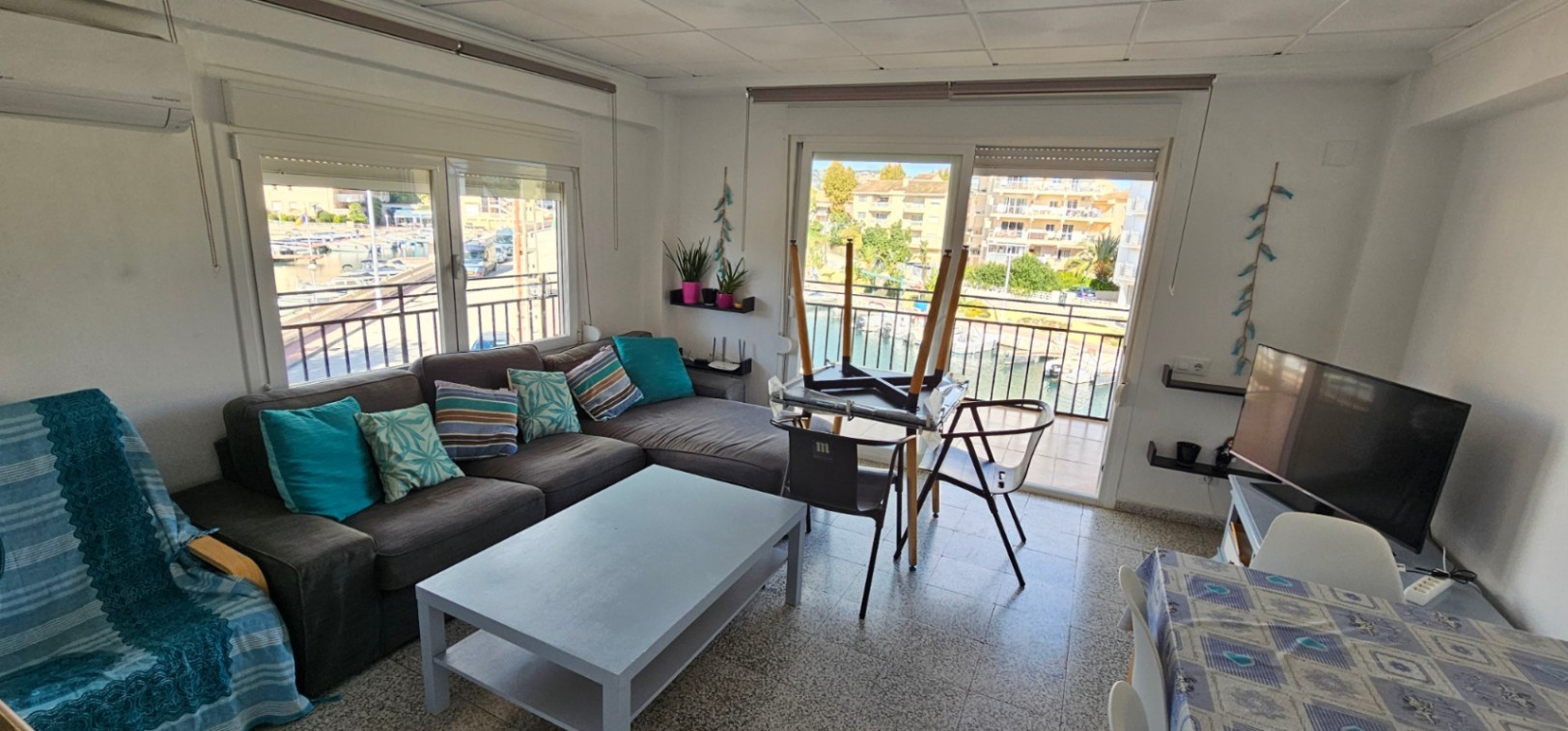 Salón luminoso con sofá rinconera, aire acondicionado y acceso directo a la terraza con vistas al canal y los barcos de la marina de Jávea.