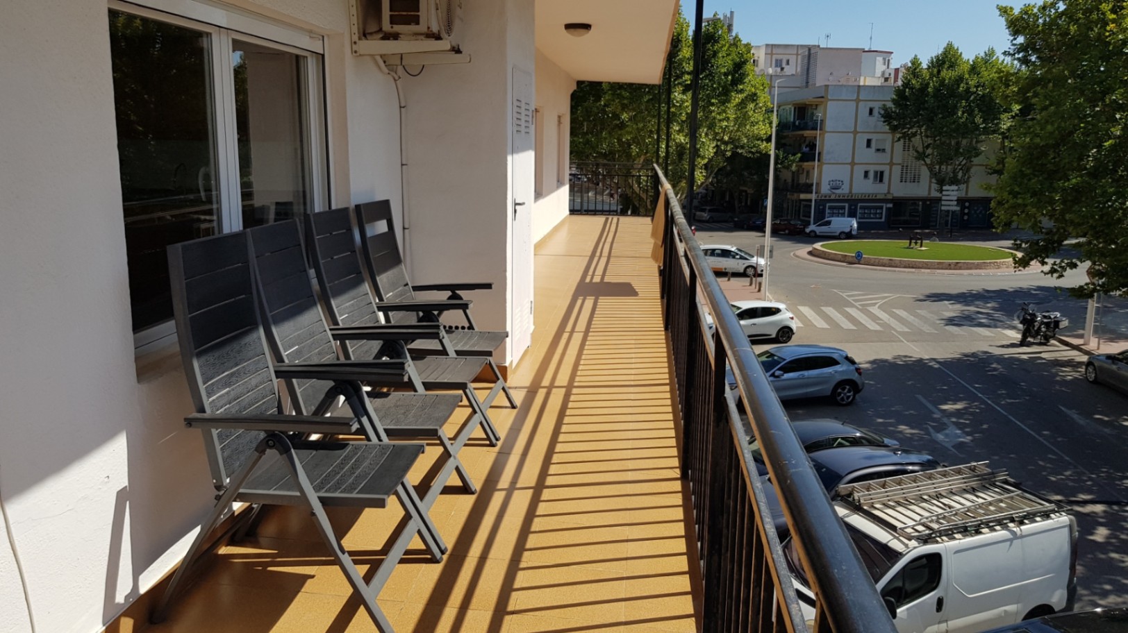 Terraza alargada con suelo cerámico ocre y barandilla metálica negra, equipada con sillas de exterior y vistas a la zona urbana arbolada de Jávea.