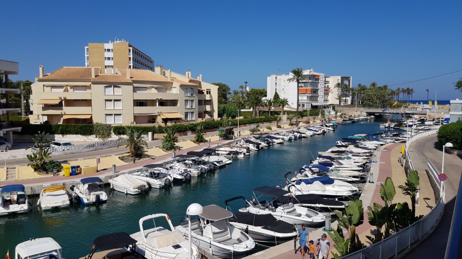 Vista desde la terraza hacia el canal de la marina con embarcaciones de recreo amarradas, rodeado de edificios residenciales y palmeras bajo un cielo despejado.