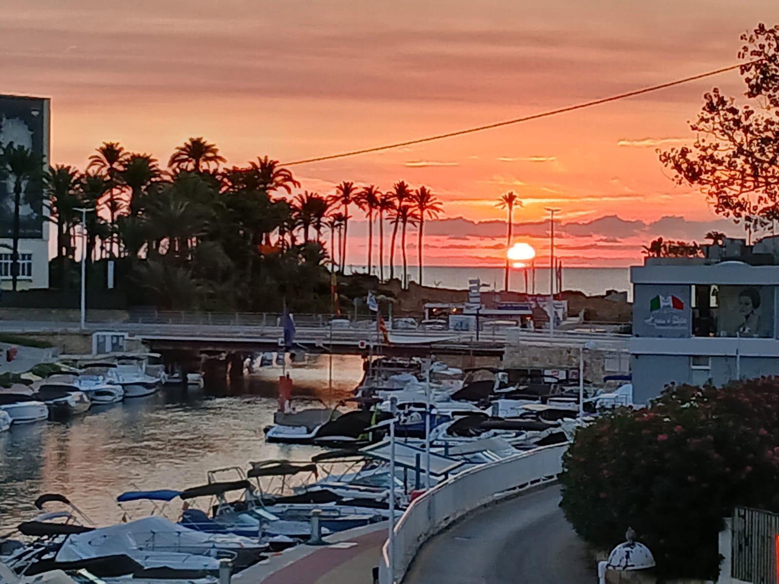 Vista panorámica desde la terraza hacia el canal de la Fontana al atardecer, mostrando los barcos amarrados y las palmeras bajo un cielo de tonos anaranjados.