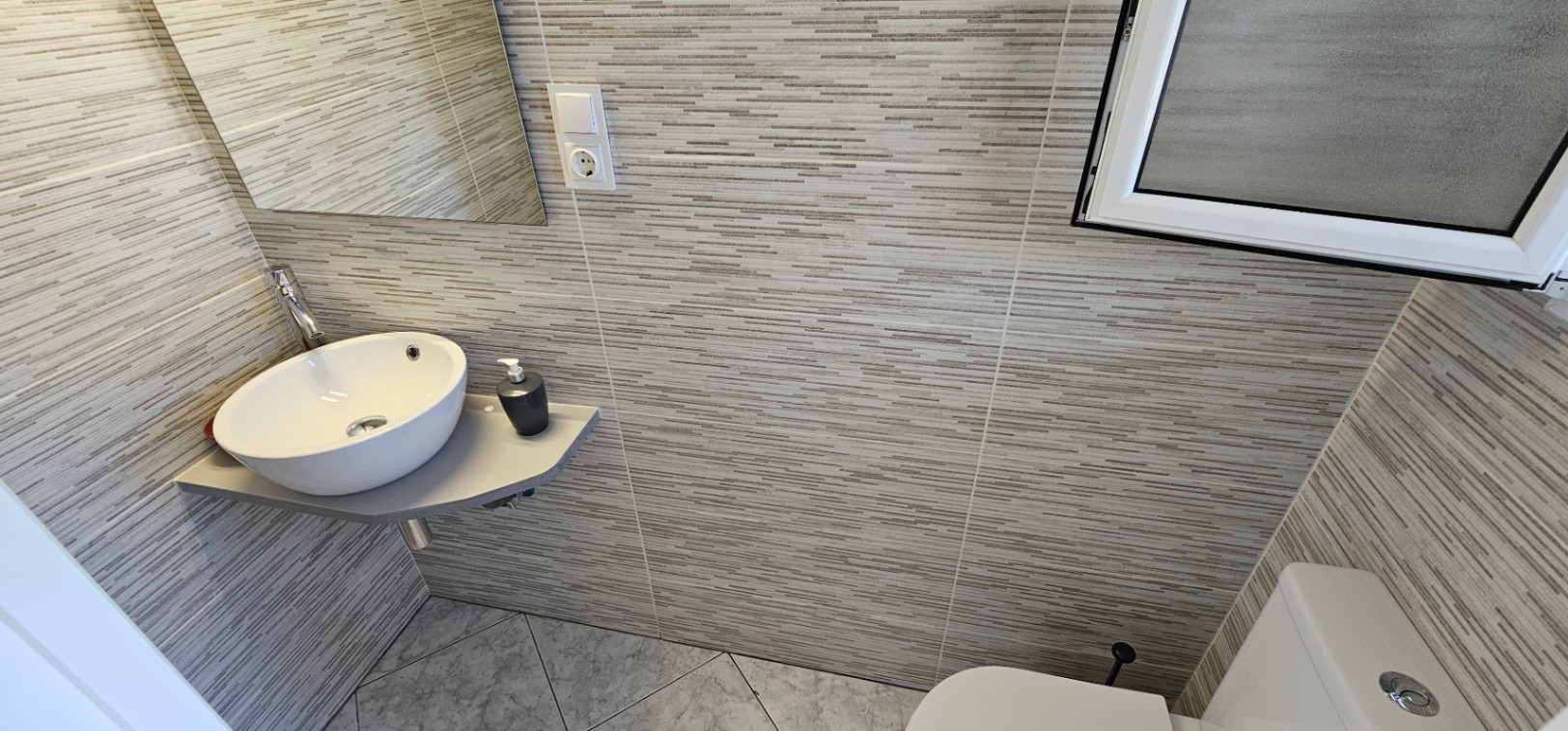 Aseo compacto con lavabo sobre encimera de esquina, paredes revestidas con azulejos de textura lineal gris y suelos cerámicos en disposición diagonal.