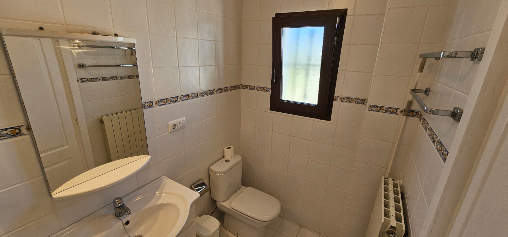 Baño funcional con paredes alicatadas en blanco y cenefa decorativa. Incluye lavabo cerámico, espejo con repisa, radiador y ventana oscurecida para mayor privacidad.
