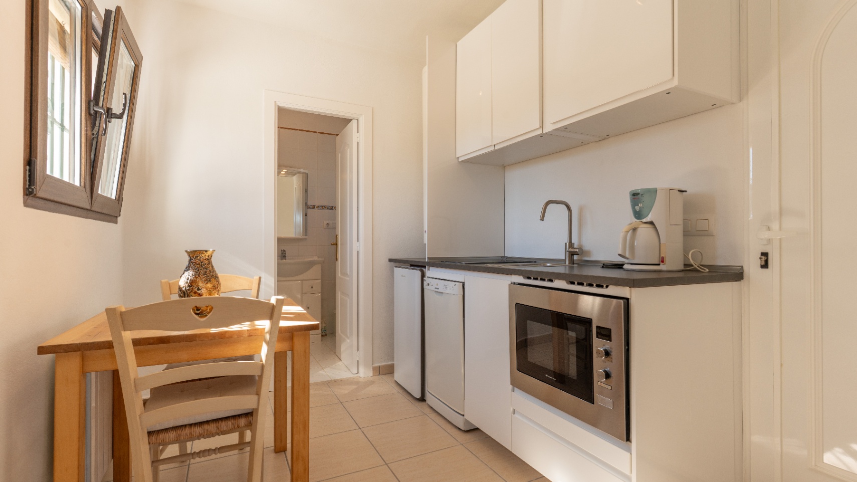 Cocina compacta con armarios blancos, electrodomésticos integrados y zona de comedor con mesa de madera, situada junto a un baño de cortesía.