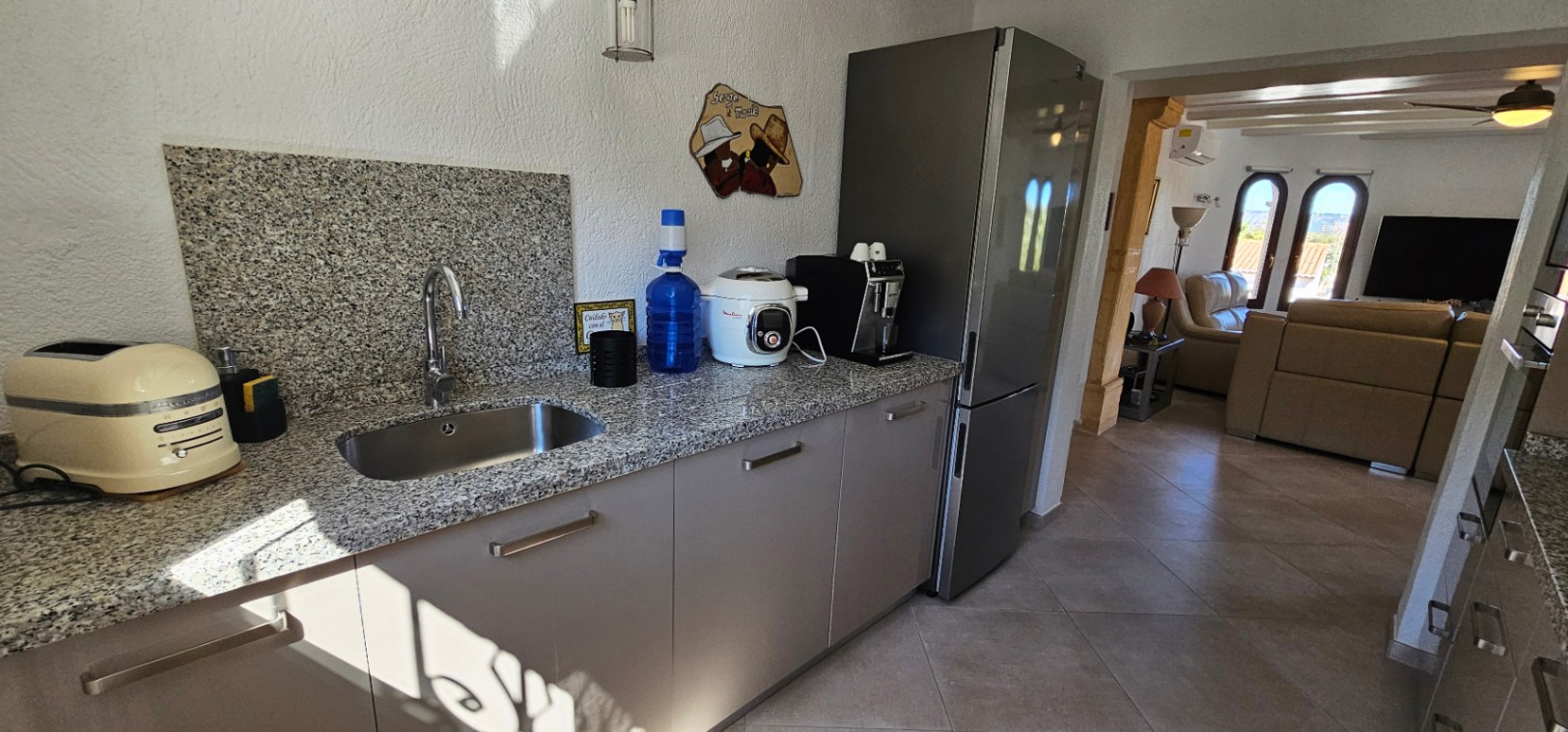 Cocina funcional con encimera de granito y fregadero de acero inoxidable, abierta hacia un salón luminoso con sofás de cuero y ventanales en arco.