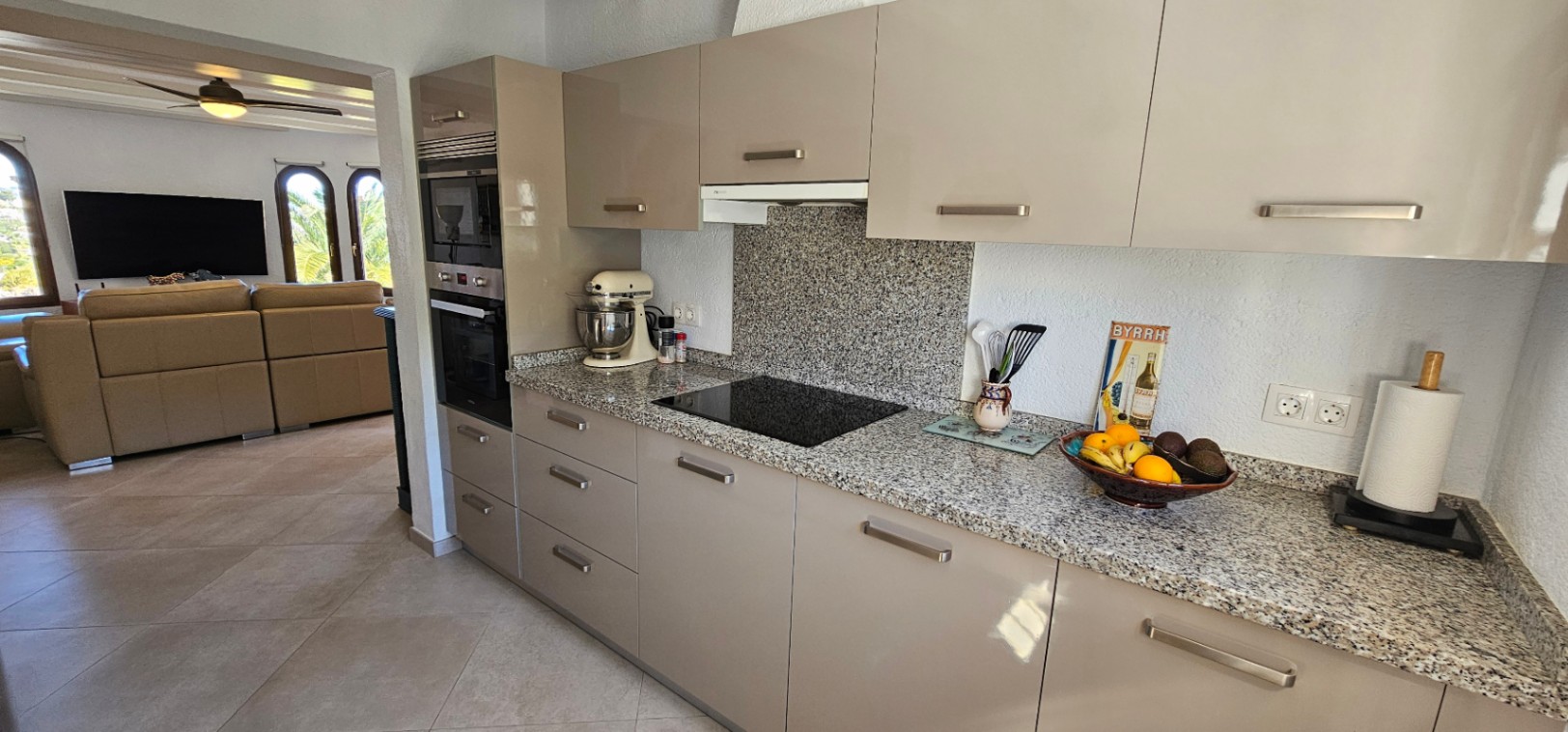 Cocina moderna con encimera de granito, armarios de alto brillo en tono visón y electrodomésticos integrados, abierta hacia un luminoso salón con ventanales de arco.