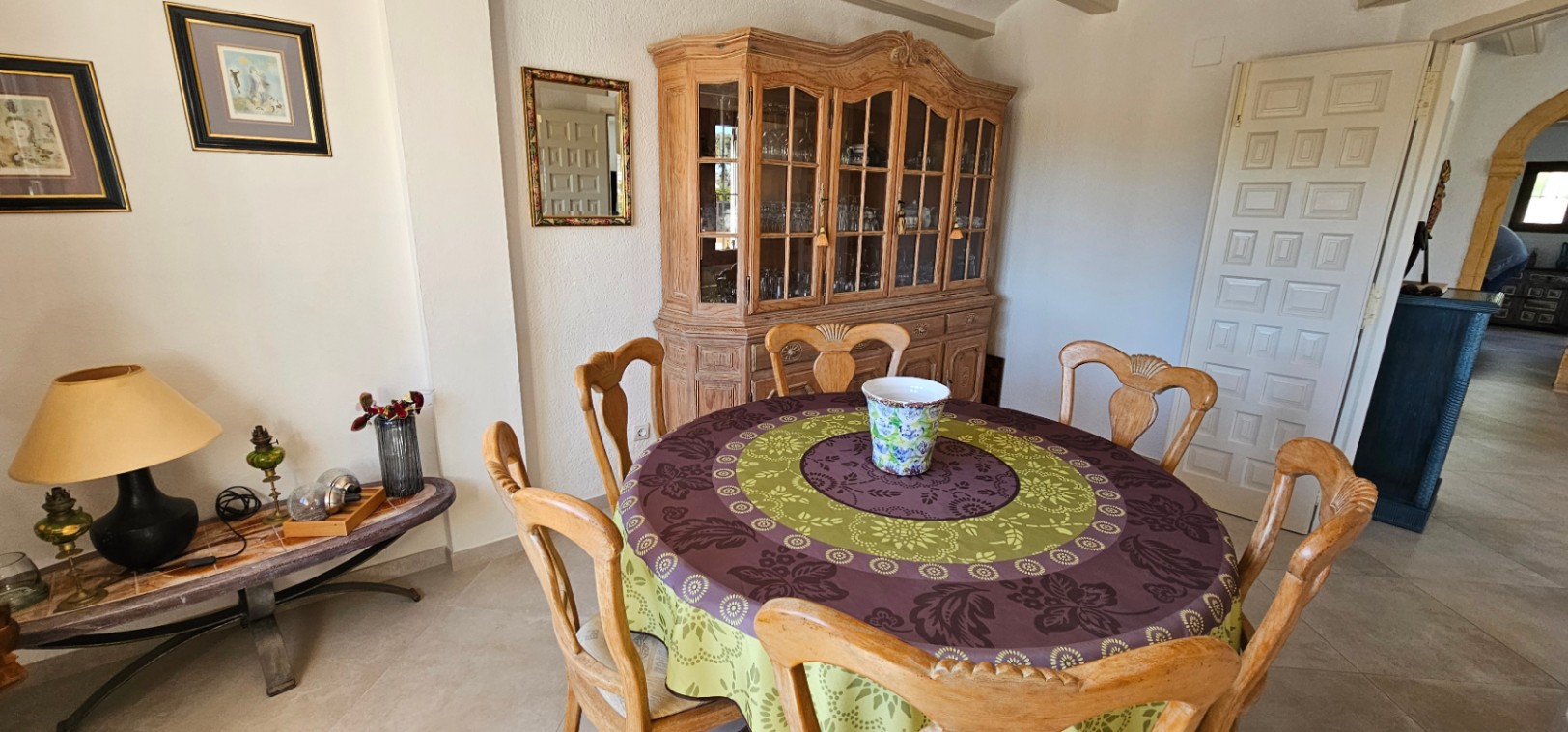 Comedor de estilo rústico con mesa redonda de madera, sillas talladas y una amplia vitrina clásica. El espacio cuenta con suelos cerámicos y paredes de tono neutro.