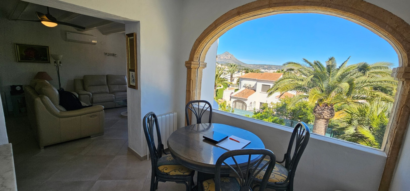 Comedor en terraza acristalada con arco de piedra tosca, ofreciendo vistas despejadas al Montgó. Conecta con un salón equipado con sofás de cuero y aire acondicionado.