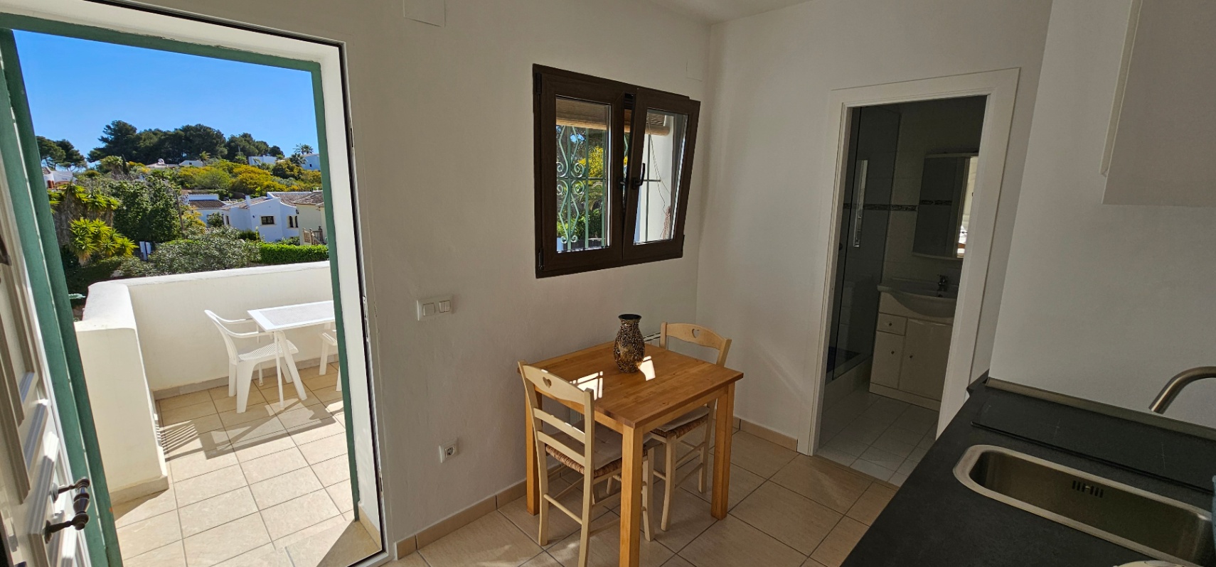 Comedor funcional con mesa de madera y sillas, conectado a una terraza exterior y un baño completo. Destaca la luz natural y el suelo cerámico uniforme.