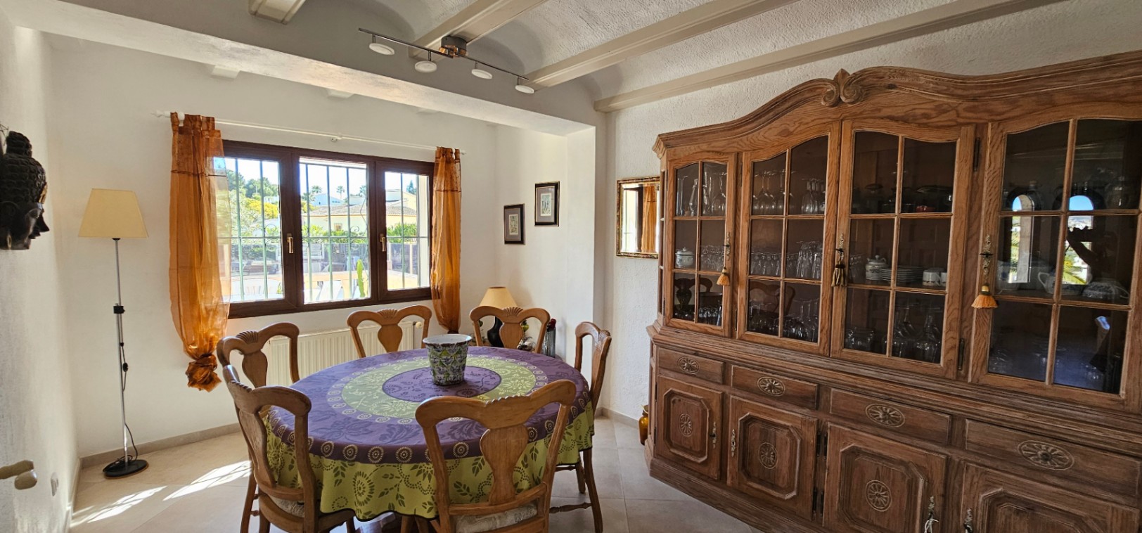 Comedor tradicional con techos de vigas vistas, gran alacena de madera tallada y mesa redonda. La ventana de madera aporta abundante luz natural y vistas al jardín.