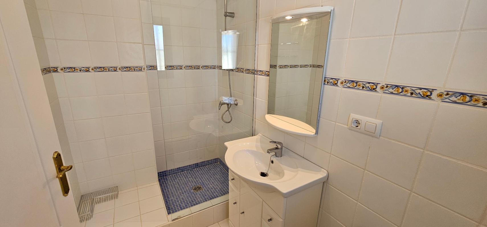 Cuarto de baño funcional con ducha de mampara de cristal, suelo de gresite azul y paredes alicatadas en blanco con una cenefa decorativa de estilo mediterráneo.