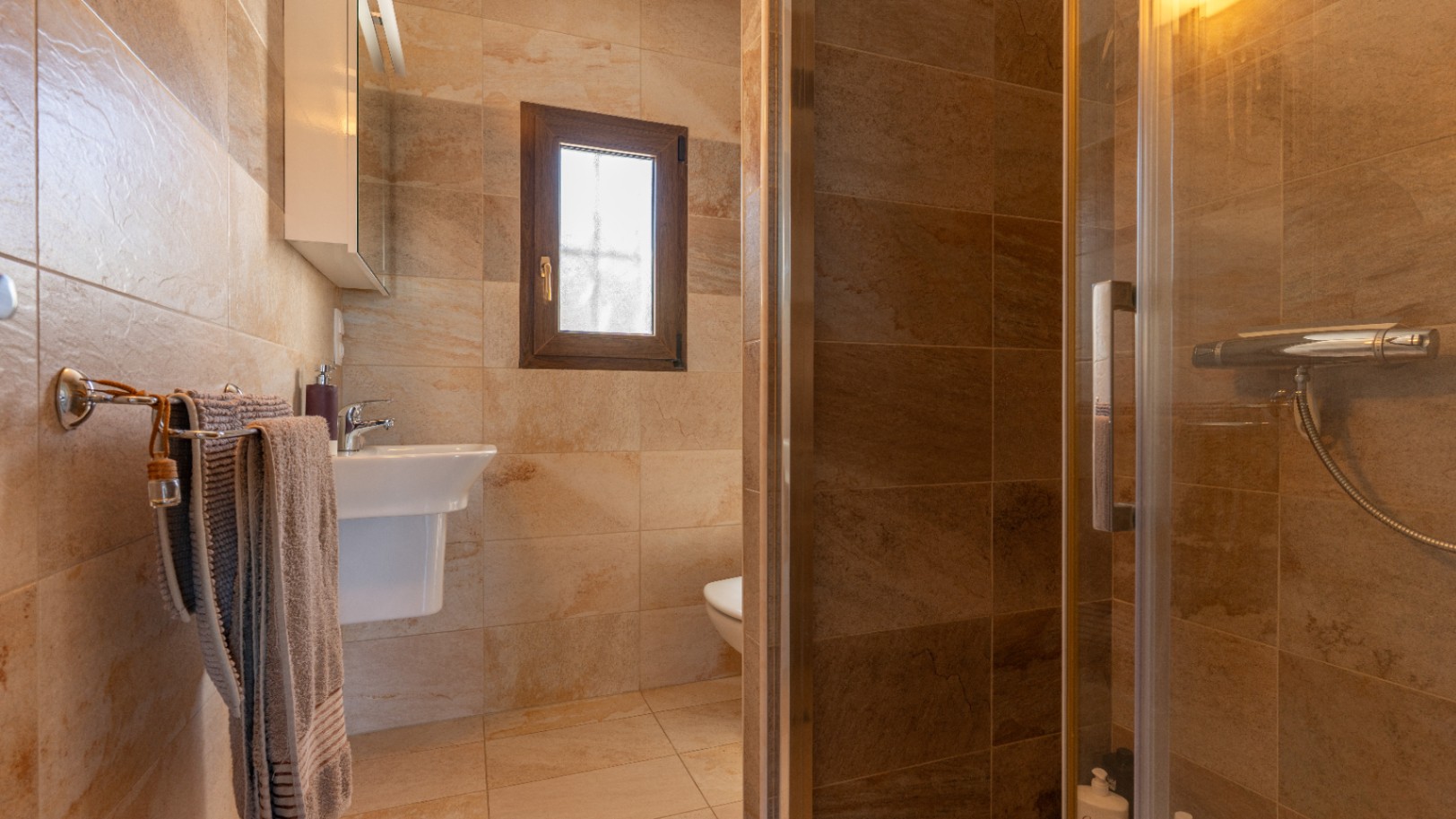 Cuarto de baño revestido en gres cerámico de tonos tierra, equipado con ducha acristalada, lavabo suspendido y ventana de madera con cristal traslúcido.