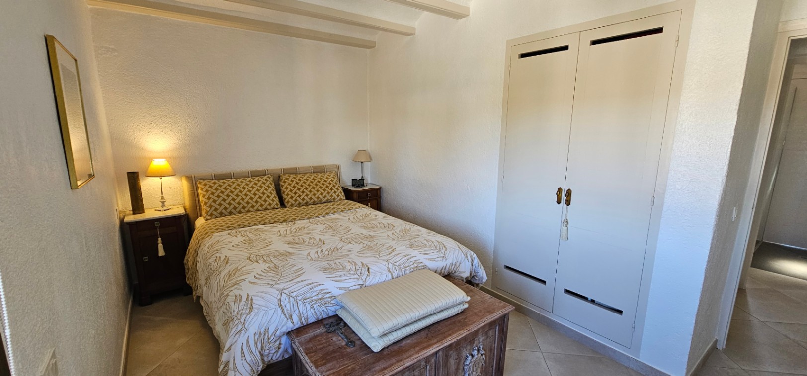 Dormitorio acogedor con vigas vistas en el techo, armario empotrado blanco y un arcón rústico de madera a los pies de la cama de matrimonio.