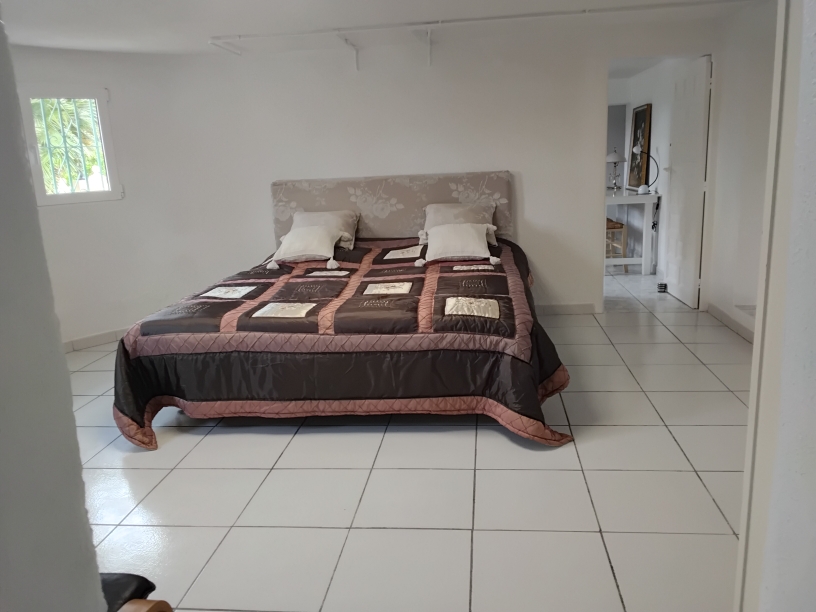 Dormitorio amplio con suelos de baldosa blanca, paredes despejadas y una cama doble con cabecero floral. Incluye una ventana con reja de seguridad y acceso a una estancia contigua.