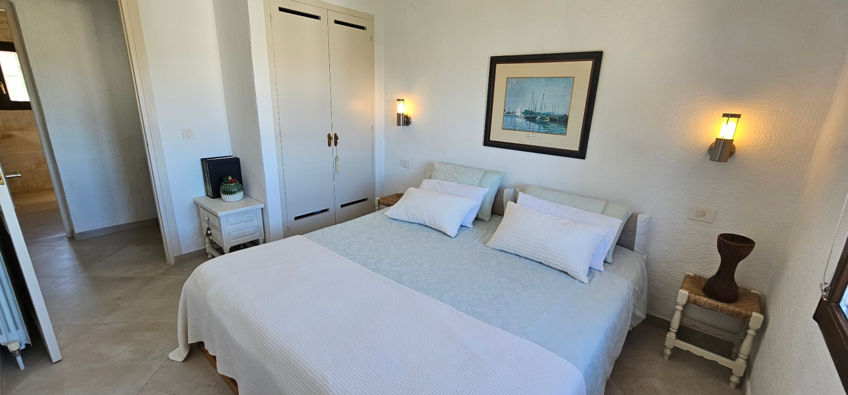 Dormitorio con cama doble, armario empotrado blanco y apliques de pared. Destacan el suelo de gres y la luz cálida que resalta las paredes texturizadas.