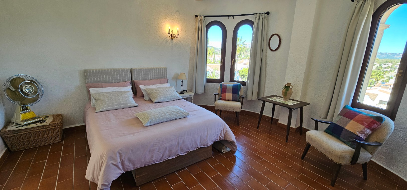 Dormitorio luminoso con suelo de terracota, ventanas de arco de madera y mobiliario de estilo rústico. Incluye zona de estar con sillones y vistas despejadas al entorno de Jávea.