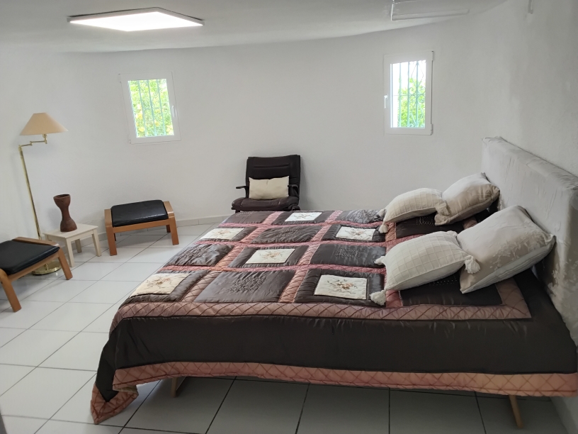 Dormitorio luminoso con suelos de baldosa blanca, cama doble con cabecero tapizado y ventanas con rejas de seguridad que permiten la entrada de luz natural.