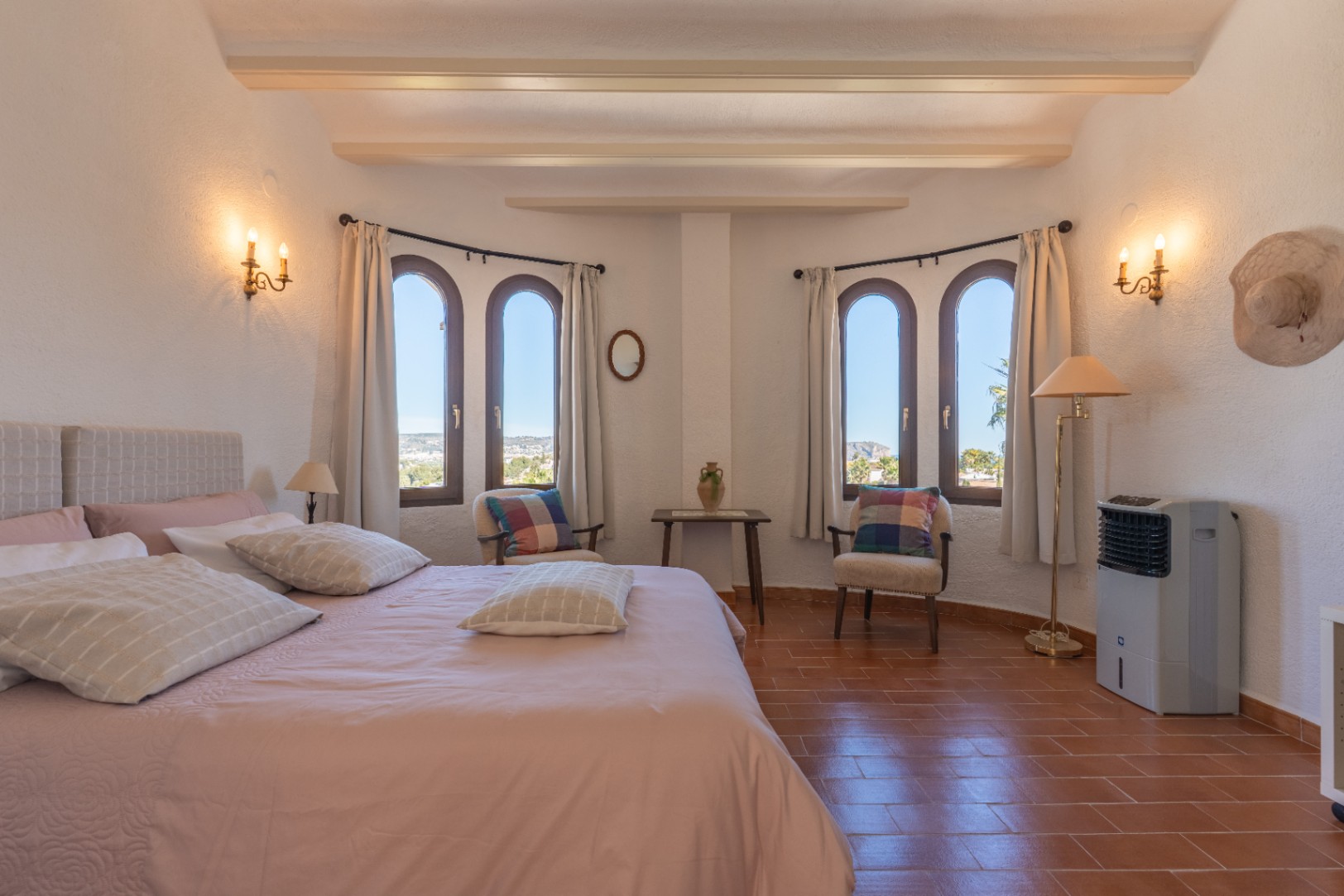 Dormitorio luminoso con suelos de terracota, vigas vistas y cuatro ventanas de arco que ofrecen vistas despejadas. Incluye zona de estar y apliques de pared clásicos.