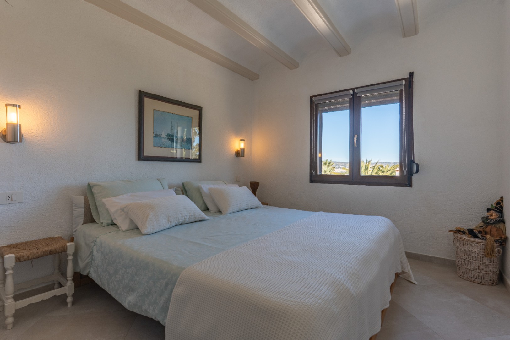 Dormitorio luminoso con vigas decorativas en el techo, apliques de pared modernos y ventana de madera con vistas al entorno natural de Jávea.