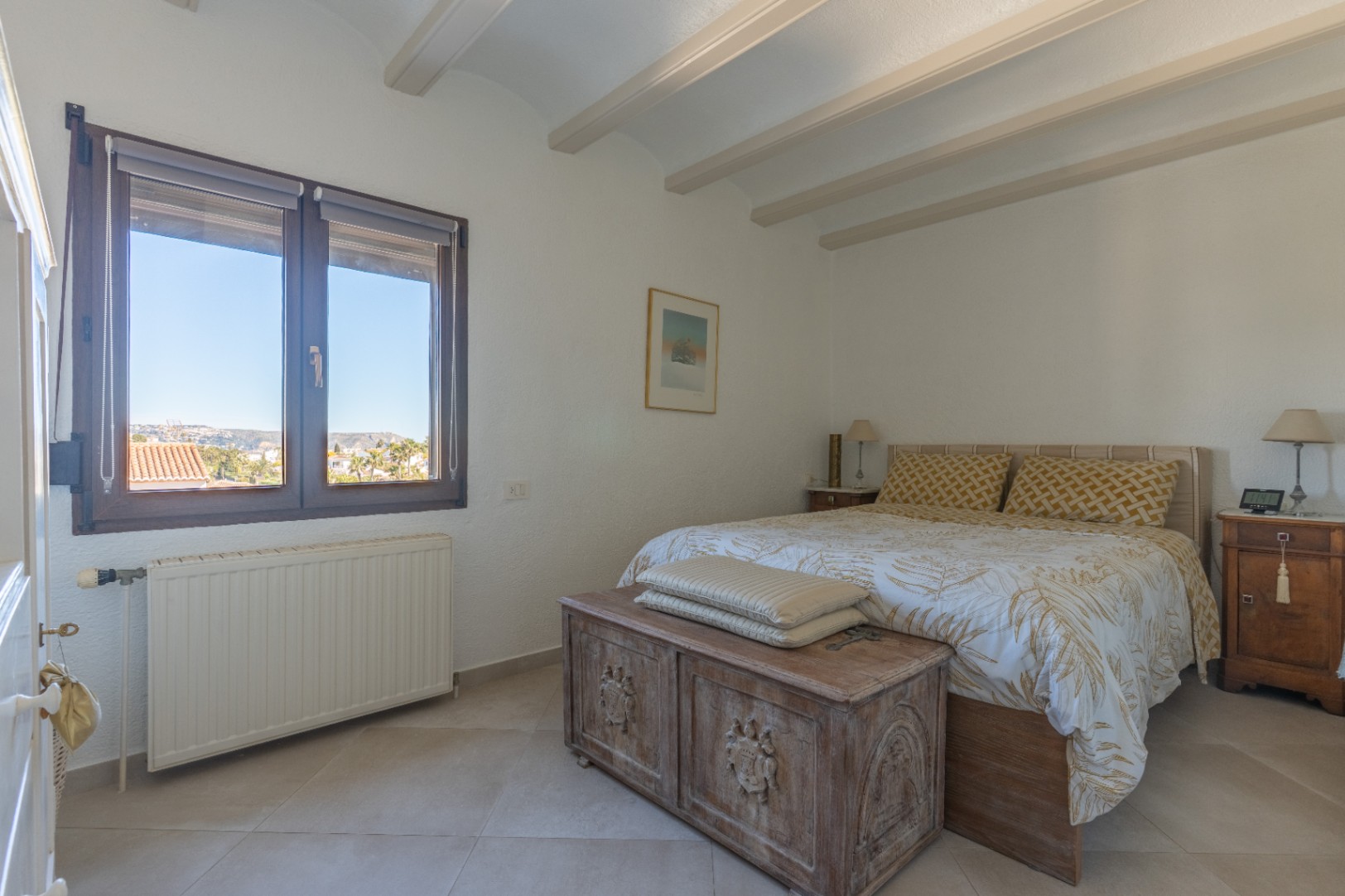 Dormitorio luminoso con vigas vistas en el techo, ventana de madera con vistas despejadas y un arcón rústico tallado a los pies de la cama.