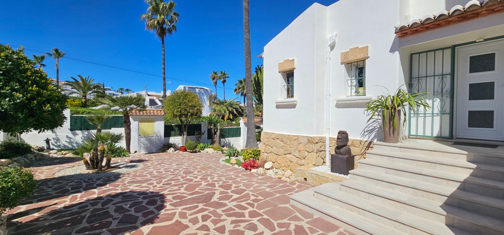Entrada principal con escalinata de piedra, fachada blanca con zócalo de piedra natural y patio pavimentado rodeado de vegetación mediterránea y palmeras.