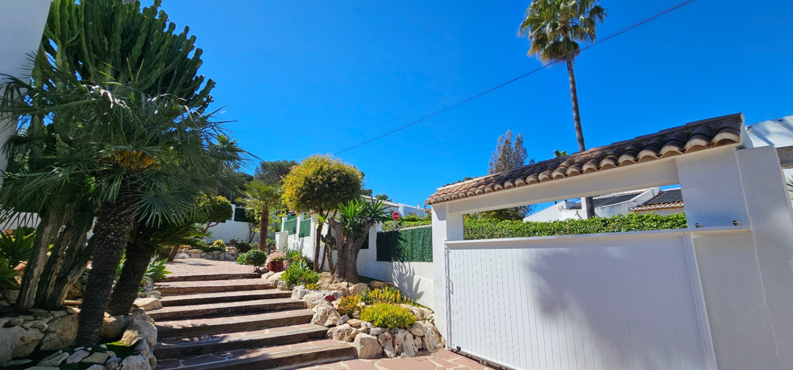 Entrada principal con puerta corredera blanca y escalinata de piedra rodeada de vegetación mediterránea, incluyendo cactus y palmeras bajo un cielo despejado.