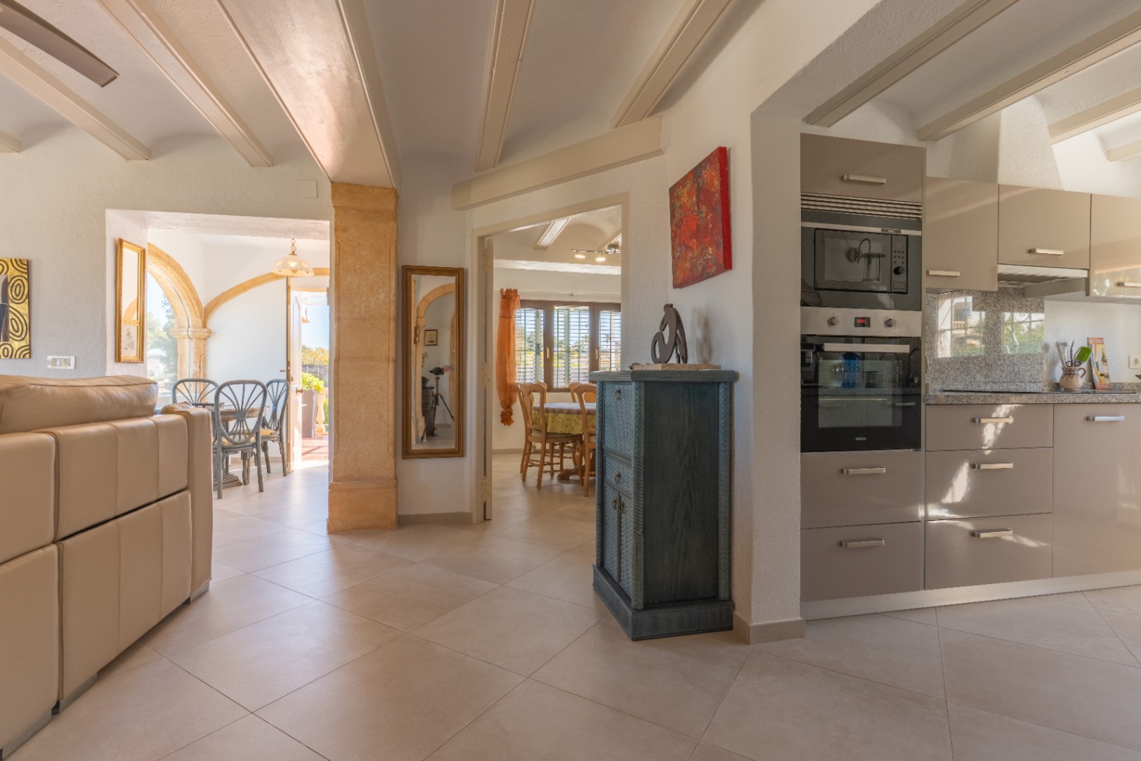 Espacio diáfano que integra cocina moderna, salón y comedor, destacando las vigas vistas en el techo y pilares de piedra Tosca tradicional.