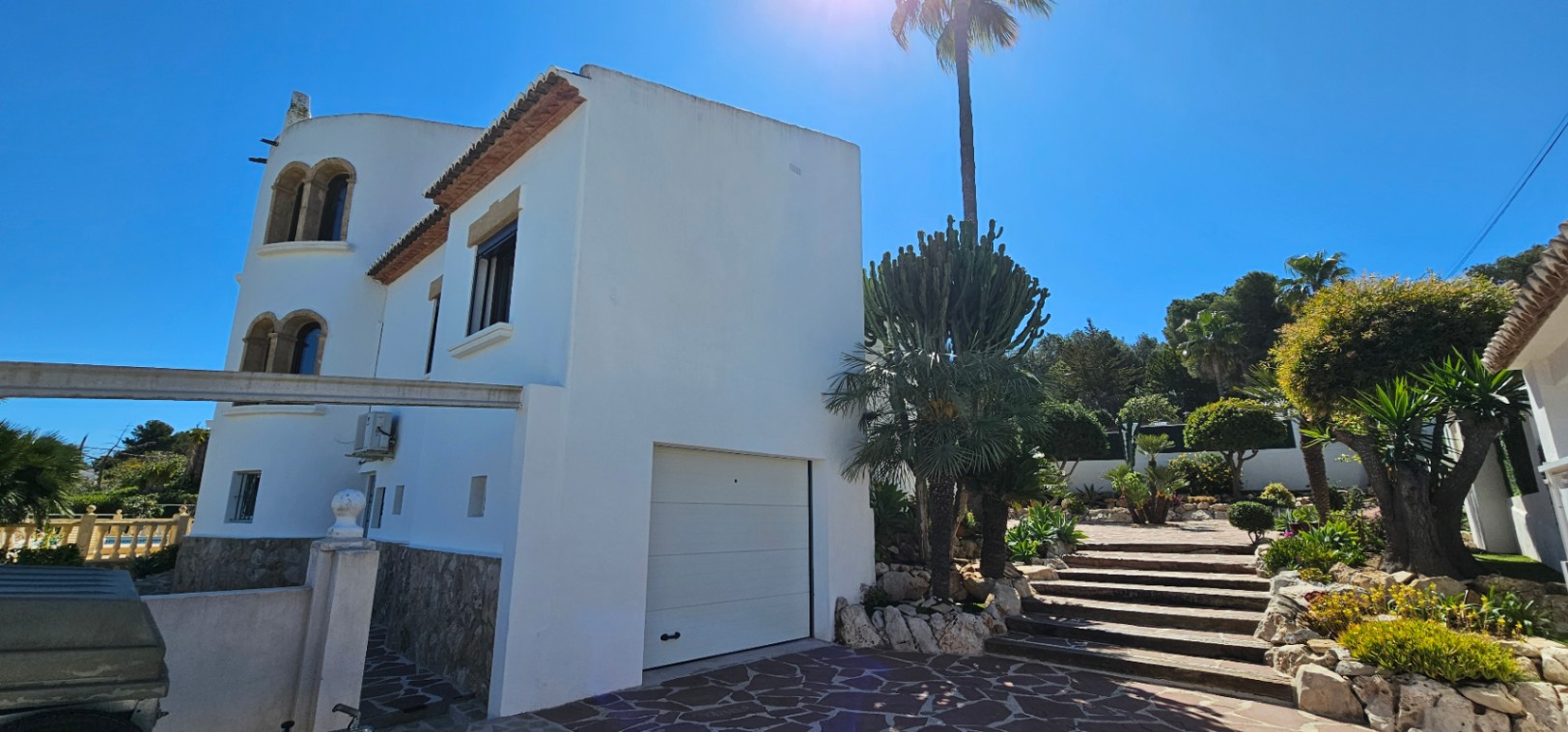 Fachada blanca con base de piedra natural, garaje integrado y escalinata hacia el jardín mediterráneo con palmeras y cactus bajo el sol de Jávea.