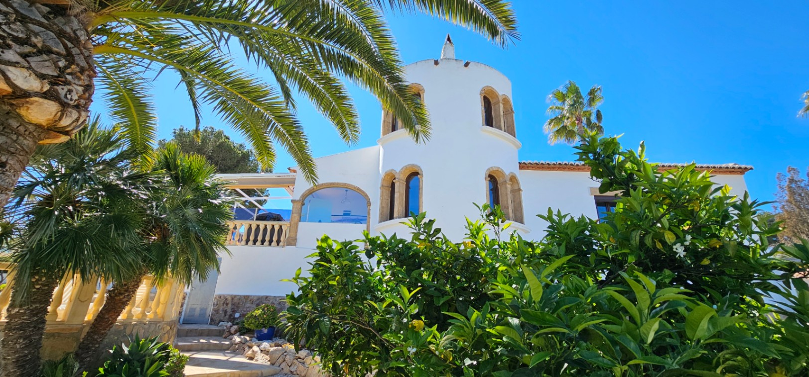 Fachada blanca de estilo mediterráneo con una torre circular, ventanas de piedra tosca y rodeada de vegetación exuberante con palmeras y cítricos bajo un cielo despejado.