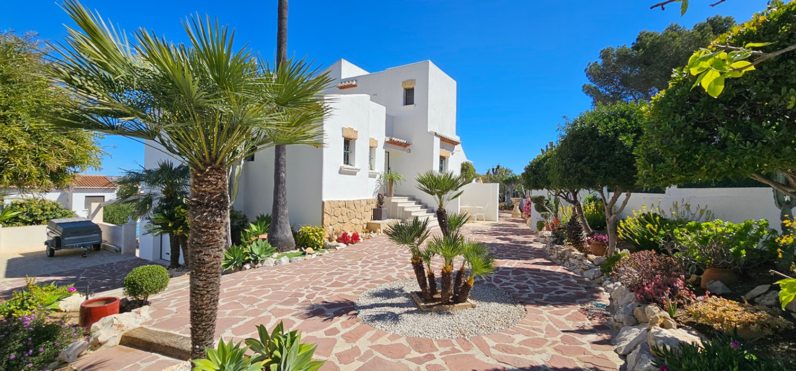 Fachada blanca de estilo mediterráneo con zócalo de piedra natural, rodeada de un jardín cuidado con palmeras y pavimentación de piedra irregular.