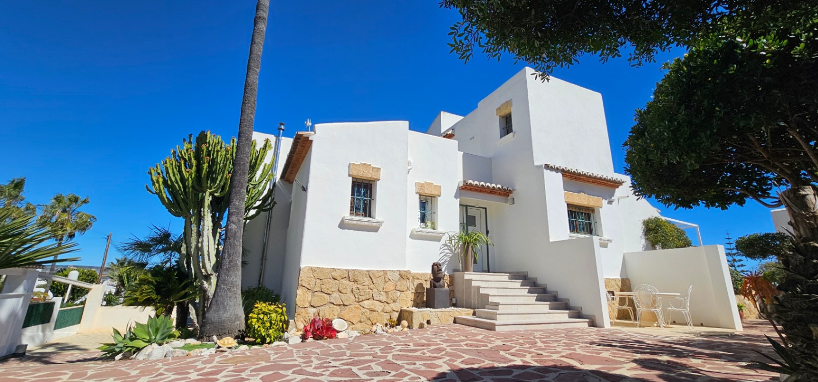 Fachada mediterránea con muros blancos, zócalo de piedra natural y acceso por escalinata. El exterior incluye un patio pavimentado y vegetación autóctona con cactus y palmeras.