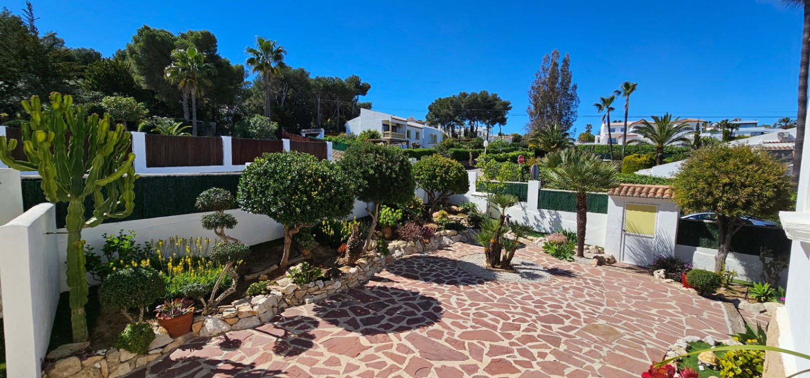Patio exterior con pavimento de piedra irregular rojiza, rodeado de muros blancos y un jardín mediterráneo con palmeras, cactus y arbustos ornamentales.