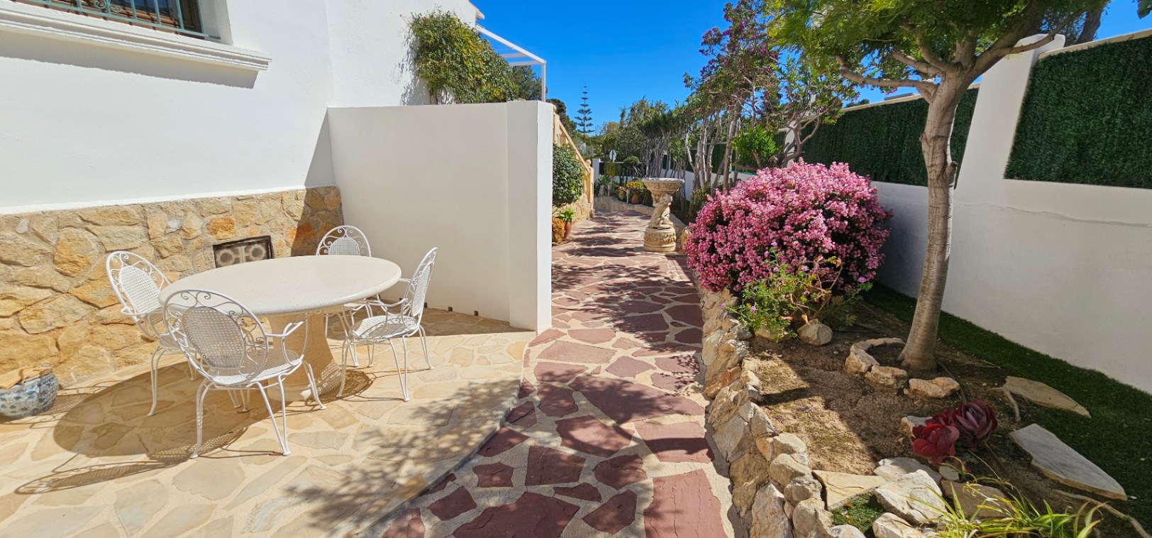 Patio exterior con pavimento de piedra natural, mobiliario de hierro forjado blanco y jardines mediterráneos con arbustos en flor y muros de piedra.