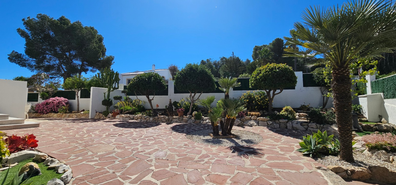 Patio exterior pavimentado con piedra natural irregular, rodeado de jardineras con palmeras, arbustos mediterráneos y muros blancos bajo un cielo despejado.