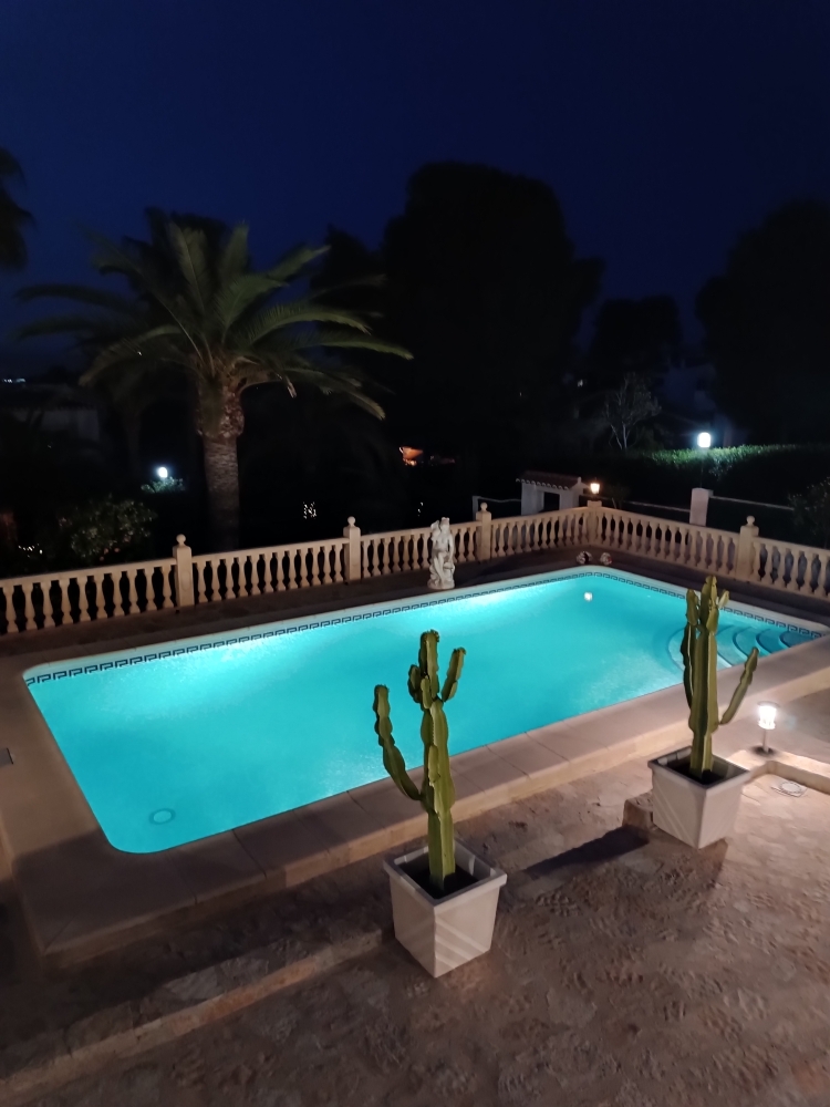 Piscina iluminada de noche con tonos turquesa, rodeada de una terraza de piedra natural con balaustrada clásica y cactus decorativos en maceteros blancos.
