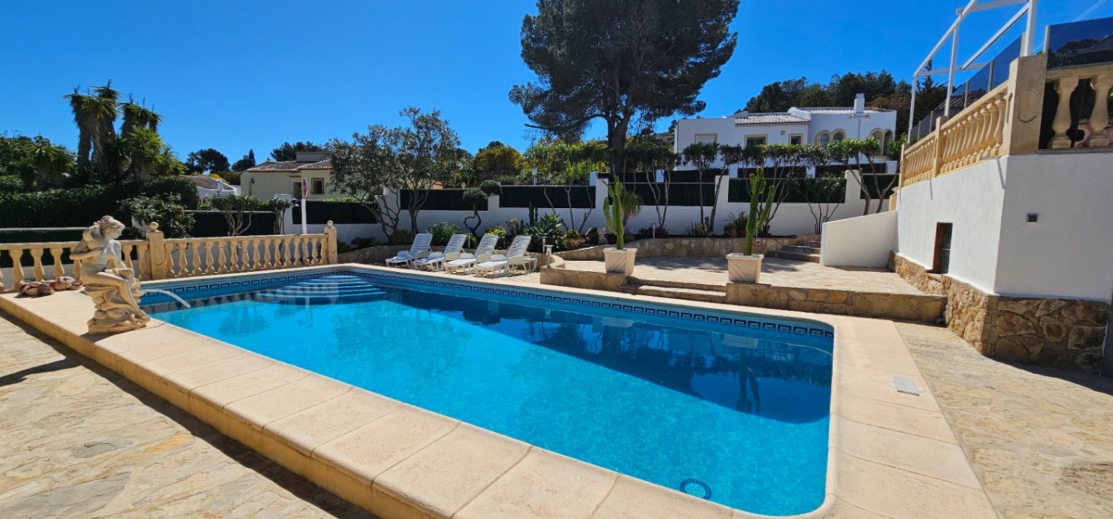 Piscina rectangular con escalera romana rodeada de una amplia terraza de piedra, zona de hamacas y jardín mediterráneo con cactus y pinos.